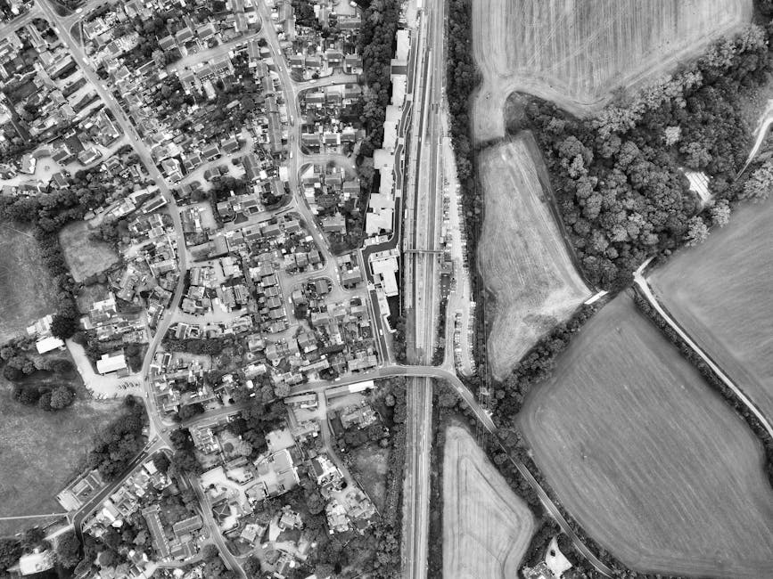 An aerial view of a rural landscape featuring large, well-maintained green fields with visible mowing lines, bordered by neatly trimmed hedges and narrow dirt paths. In the background, there is a smal